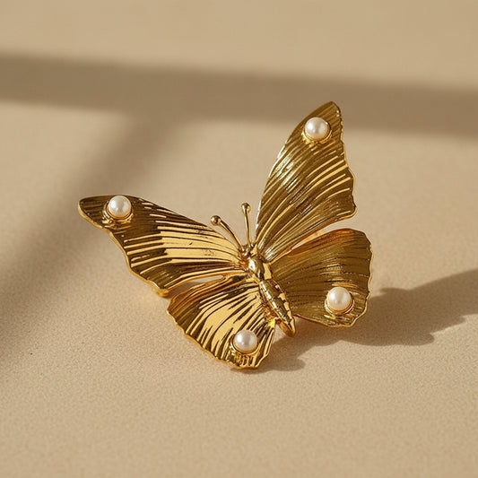 Exquisite gold butterfly brooch with finely textured radiating wings and small pearl accents, displayed on a warm beige surface with artistic sunlight shadows.