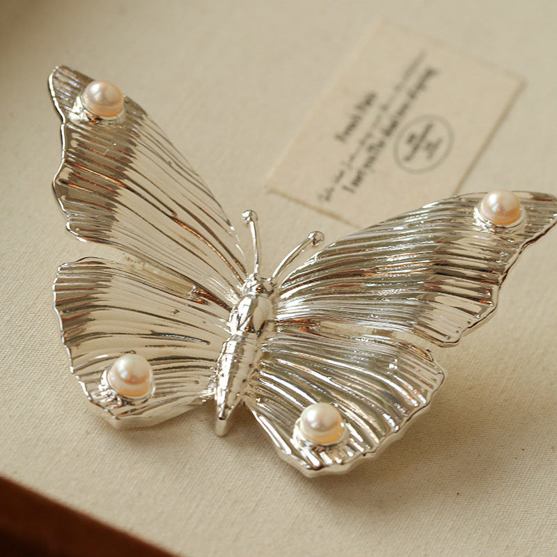 Close-up of the silver butterfly brooch variant, showcasing the intricate metallic engraving and the luminous pearls on the wingtips.