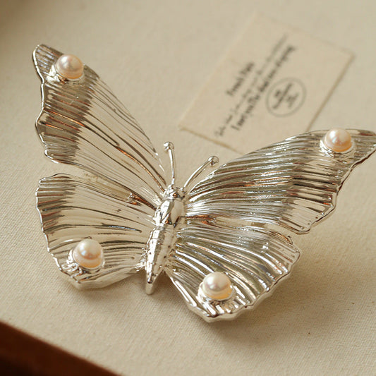 Close-up of the silver butterfly brooch variant, showcasing the intricate metallic engraving and the luminous pearls on the wingtips.