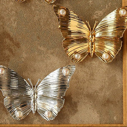 Top view of both gold and silver butterfly brooches side-by-side on a rustic background, highlighting the contrast between the two metallic finishes.
