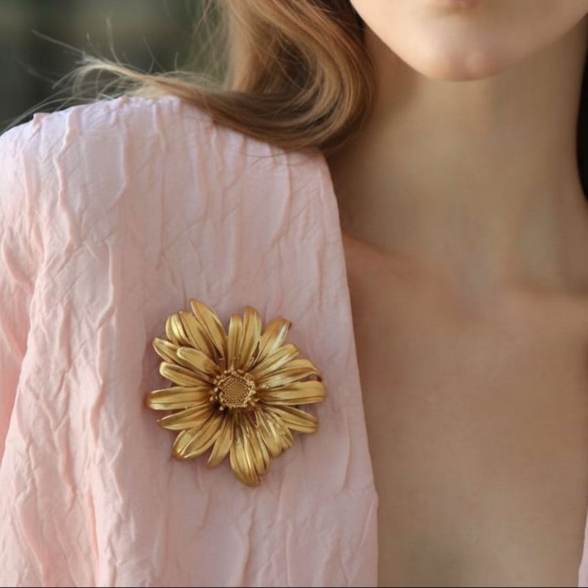 Lifestyle shot of a woman wearing the statement gold flower brooch on a light pink textured blazer, demonstrating its scale and luxury aesthetic.