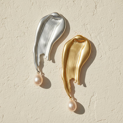 A pair of organic, brush-stroke shaped metal brooches in brushed silver and matte gold, with a baroque drop pearl dangling from the base of each, resting on a textured cream surface.