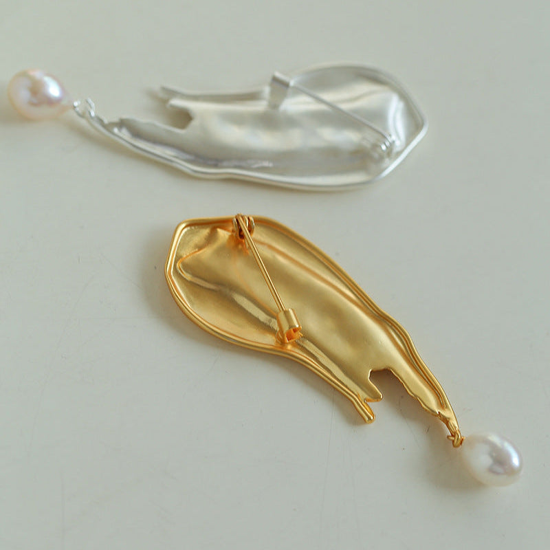 Overhead view of the silver and gold fluid-form brooches together on a neutral light background, displaying the full color options and pearl placement.