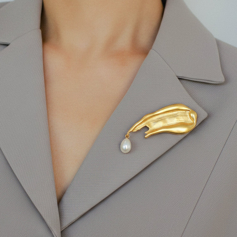 Lifestyle image of a model wearing the matte gold, artful brush-stroke brooch with a hanging pearl on a grey structured blazer, showing the piece's sophisticated size and wearability.