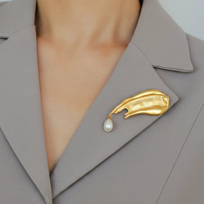Lifestyle image of a model wearing the matte gold, artful brush-stroke brooch with a hanging pearl on a grey structured blazer, showing the piece's sophisticated size and wearability.