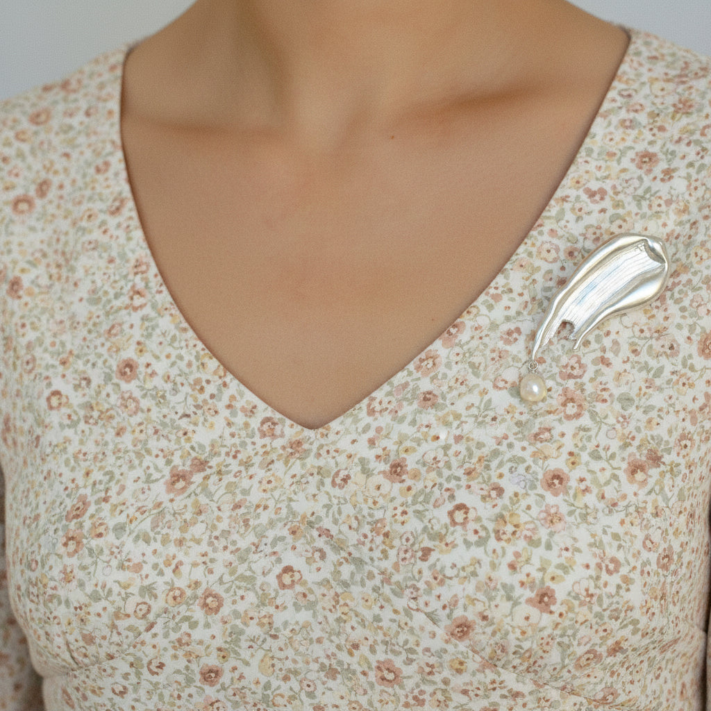 A view of a model in a v-neck floral top wearing the silver, liquid-metal style brooch with a pearl dangle, demonstrating how the piece adds an artistic touch to a casual outfit.