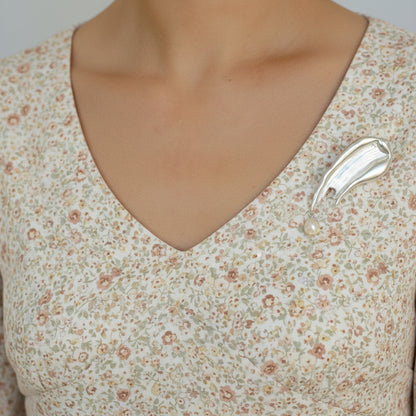 A view of a model in a v-neck floral top wearing the silver, liquid-metal style brooch with a pearl dangle, demonstrating how the piece adds an artistic touch to a casual outfit.