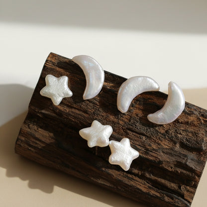 Artistic set of crescent moon and star-shaped stud earrings made from iridescent mother-of-pearl, displayed on a piece of dark textured wood in warm natural sunlight.