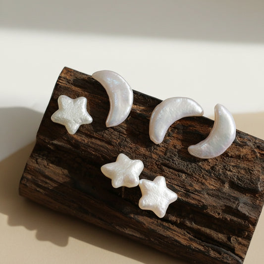 Artistic set of crescent moon and star-shaped stud earrings made from iridescent mother-of-pearl, displayed on a piece of dark textured wood in warm natural sunlight.