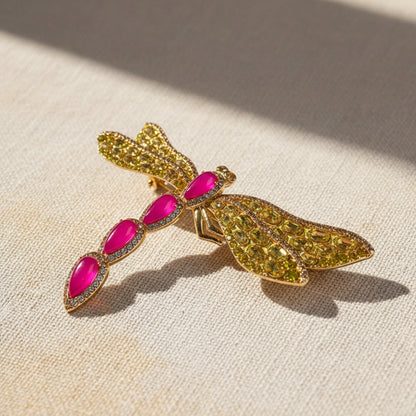 High-end gold-plated brass dragonfly brooch, intricately set with fuchsia cold enamel and honey-yellow pavé crystals. A large statement jewelry piece for women, displayed against a rustic linen background with soft, natural sunlight and shadows.