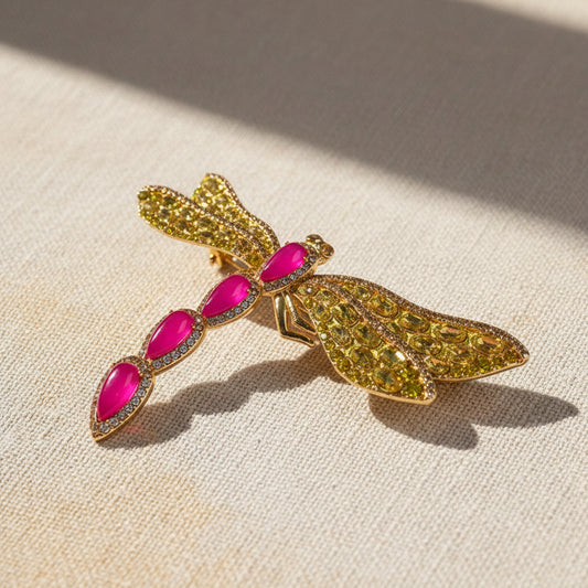 High-end gold-plated brass dragonfly brooch, intricately set with fuchsia cold enamel and honey-yellow pavé crystals. A large statement jewelry piece for women, displayed against a rustic linen background with soft, natural sunlight and shadows.