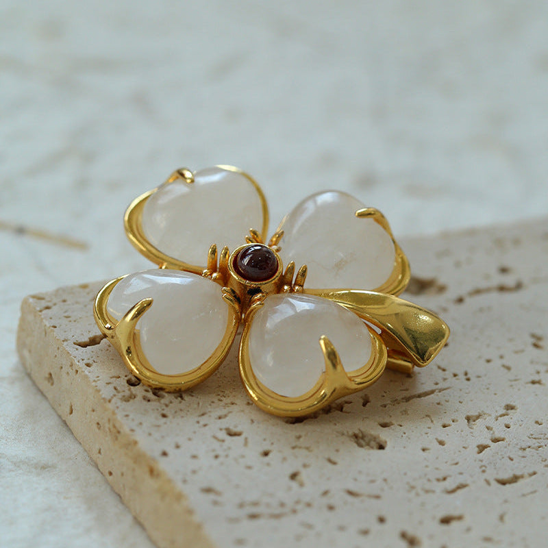 Vintage-style four-petal flower brooch featuring translucent white stone petals with gold-plated edges and a small red garnet-like center, resting on a textured stone base.