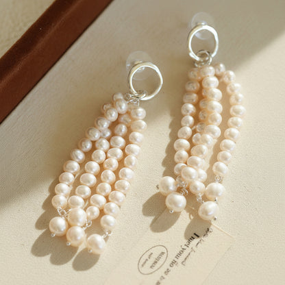 Dramatic multi-strand pearl tassel earrings with silver circular studs, resting on a warm neutral surface with elegant natural sunlight and shadows.